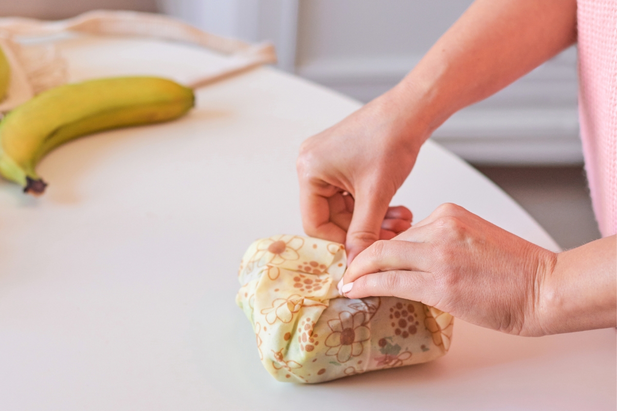 cropped photo of woman wrapping a sandwich in a bees wax wrap with fruit in background